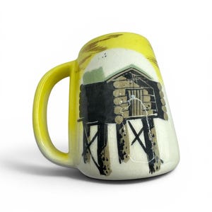 May include: A yellow ceramic mug with a curved handle. The mug features a hand-painted illustration of a cabin on stilts, with a yellow sky and black and brown details. The mug has a glossy finish.