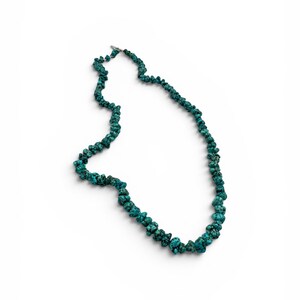 Puede incluir: Un collar compuesto por numerosas cuentas de turquesa peque&ntilde;as, irregulares y pulidas, en tonos azul verdoso y verde azulado profundo. Las cuentas est&aacute;n ensartadas en una hebra continua, con un peque&ntilde;o cierre de metal plateado en la parte superior.