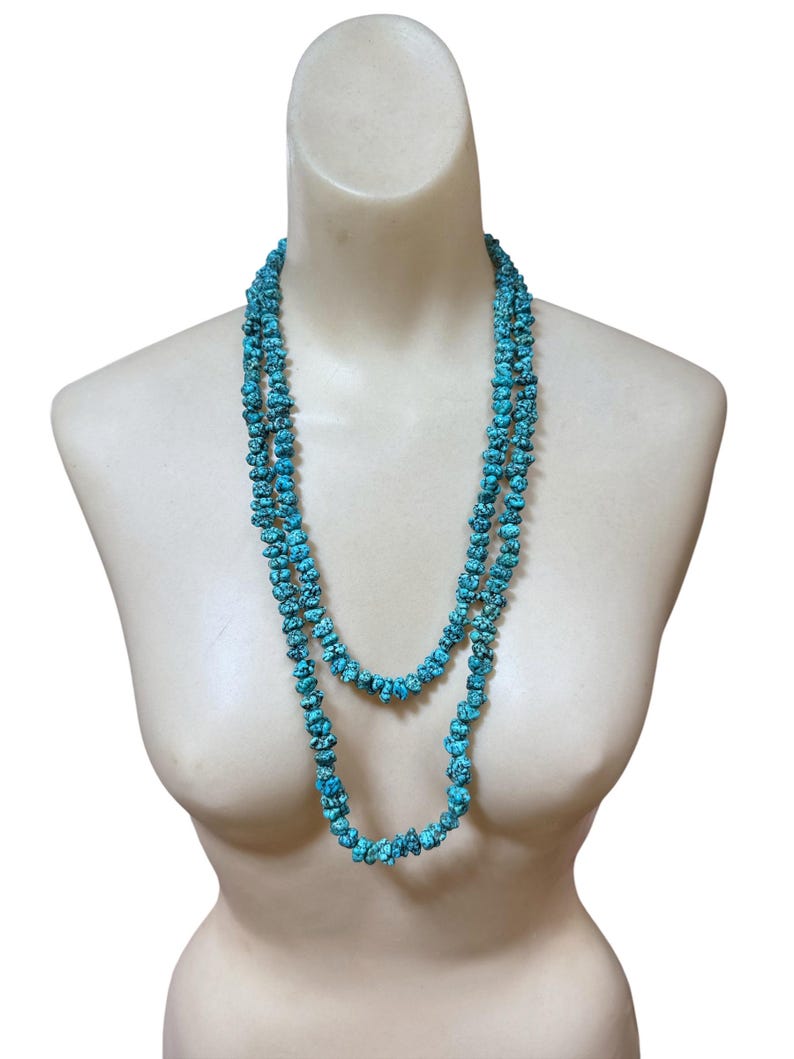 Puede incluir: Un collar largo de doble hebra que presenta numerosas piedras peque&ntilde;as, irregulares y pulidas de color turquesa con vetas de matriz oscura. Las cuentas est&aacute;n ensartadas en un bucle continuo, creando una apariencia de capas alrededor del cuello.