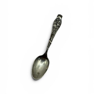 May include: A silver-toned metal spoon featuring an ornate handle with a raised relief design of a figure standing atop a globe. The handle displays intricate scrollwork patterns and a textured finish, leading to a smooth, oval-shaped bowl.