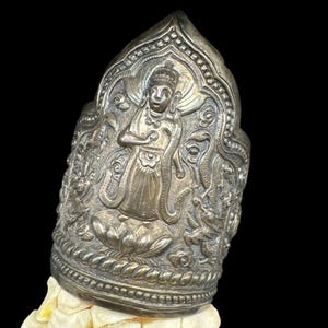 May include: A silver-colored, ornate bracelet with a detailed relief of a figure, possibly a deity, surrounded by intricate patterns. The bracelet is resting on a light-colored surface against a black background. The design suggests a cultural or religious artifact.
