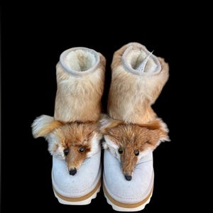 Op de afbeelding: Een paar novelty slippers met een vossendesign. De slippers hebben een lichtbruine vacht bovenkant en een witte suède voetbed. Het vossengezicht is gedetailleerd met bruine en zwarte accenten. De zolen zijn lichtbruin.
