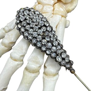 May include: A silver-toned, elongated brooch densely covered in small, clear rhinestones. The brooch is held by a skeletal hand, with a pin extending from the back. The brooch has a vintage aesthetic.