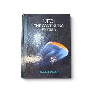 Może przedstawiać: Książka w twardej oprawie zatytułowana UFO: THE CONTINUING ENIGMA wydawnictwa Reader's Digest. Czarna okładka zawiera grafikę świecącego, niebiesko-pomarańczowego niezidentyfikowanego obiektu latającego na ciemnym, gwiaździstym tle z białym i turkusowym tekstem.