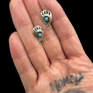 May include: A pair of silver-toned earrings, each featuring a bear paw design with a small, round turquoise stone at the center. The earrings have a small loop at the bottom for attachment. The background is black.