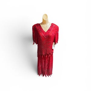 May include: A red beaded dress with a V-neckline and short sleeves. The dress features intricate beadwork and a unique, fringed hemline. The dress is displayed on a mannequin against a white background.