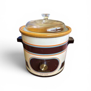 Op de afbeelding: Vintage Crock-Pot slowcooker met een heldere glazen deksel en houten afwerking. De crèmekleurige pot heeft bruine en oranje strepen en een bedieningsknop. De tekst "Crock-Pot" en "Slow-Cooker" is zichtbaar aan de voorkant.