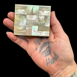 May include: A square, vintage compact mirror with a mosaic design made of iridescent mother-of-pearl tiles. The compact has a gold-colored metal frame and is held in a hand against a black background.