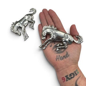Puede incluir: Dos emblemas de metal plateado, cada uno con forma de caballo encabritado. Las figuras de los caballos presentan crines, colas y patas musculosas detalladas en una pose dinámica y saltarina. La superficie metálica es lisa y reflectante.