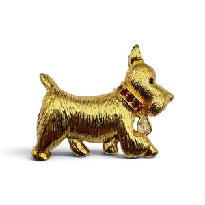 Op de afbeelding: Een goudkleurige metalen broche in de vorm van een Scottish Terrier. De hond heeft een textuurafwerking, een halsband bezet met kleine rode ronde steentjes en een enkele heldere druppelvormige kristallen hanger aan de halsband.