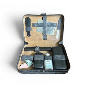 Pode incluir: Um kit de viagem vintage em couro preto aberto, revelando ferramentas de higiene pessoal. O kit contém uma navalha, uma faca pequena e frascos. O kit é fechado com um fecho preto. O conteúdo é visível.