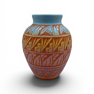 May include: A ceramic vase with a round body and a light blue rim. The vase features carved patterns in orange, yellow, and purple. The design includes geometric and stylized leaf-like motifs, creating a textured surface. The vase is approximately 15 cm tall.