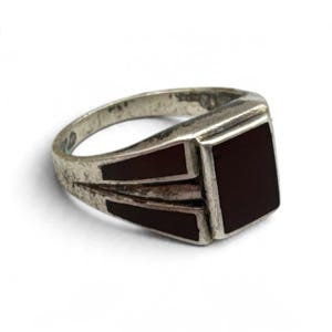 May include: A silver-toned ring featuring a rectangular dark red stone inlay on the face, with two additional trapezoidal dark red stone inlays set into the band on either side of the central setting, creating a geometric design.