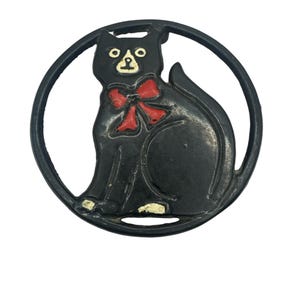 May include: A circular black metal trivet featuring a stylised cat silhouette in the centre. The cat is painted black with a bright red bow tie around its neck and white accents on its face and paws, enclosed within a solid black circular frame.
