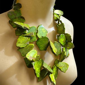 May include: A vibrant green statement necklace composed of irregularly shaped, textured stones. The necklace is displayed on a mannequin, showcasing the bold, organic design. The stones have a mottled appearance with variations in color, adding depth and visual interest.