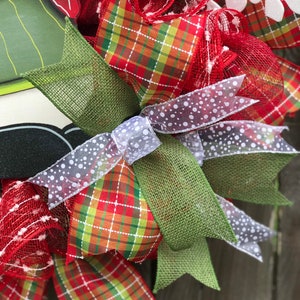 May include: A Christmas wreath with red, green, and white plaid ribbon, white mesh, and a white snowflake. The wreath is decorated with a green burlap bow and a white ribbon with white polka dots.