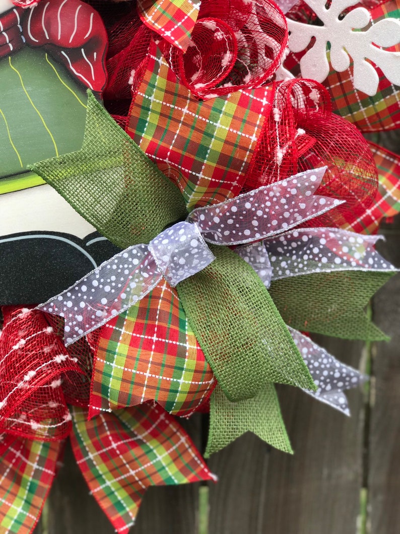 May include: A Christmas wreath with red, green, and white plaid ribbon, white mesh, and a white snowflake. The wreath is decorated with a green burlap bow and a white ribbon with white polka dots.
