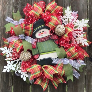 May include: A Christmas wreath with a snowman in the center. The wreath is decorated with red and green plaid ribbon, white snowflakes, and burlap balls.
