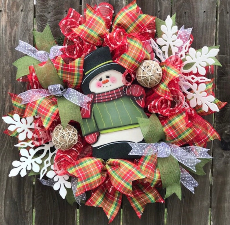 May include: A Christmas wreath with a snowman in the center. The wreath is decorated with red and green plaid ribbon, white snowflakes, and burlap balls.