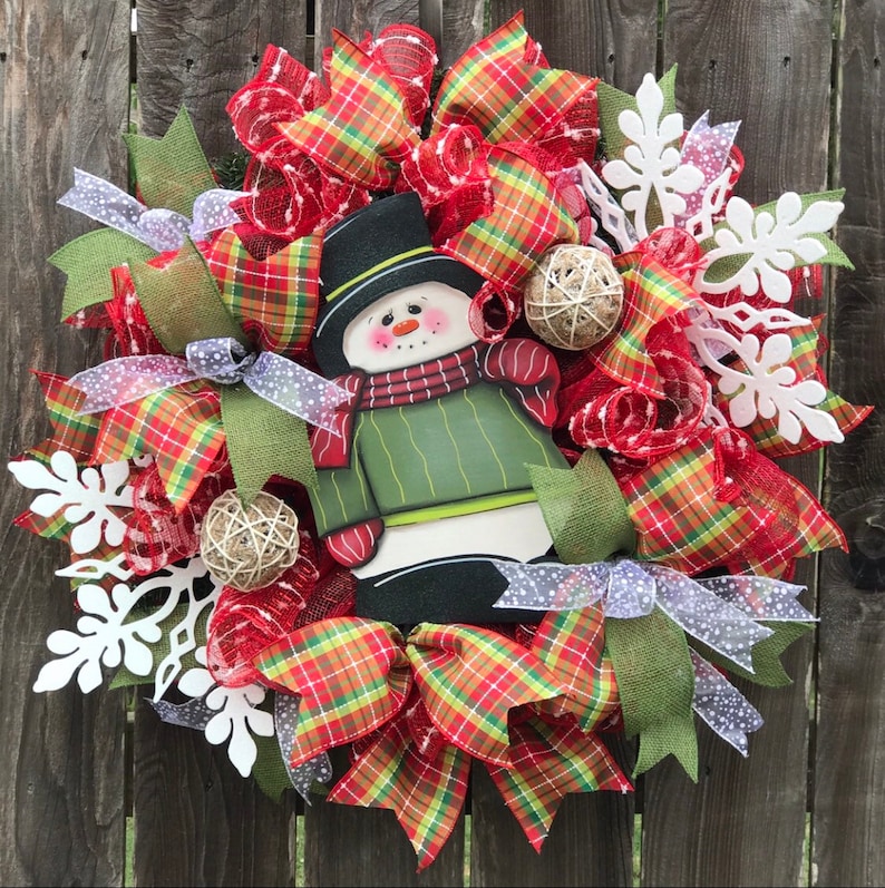 May include: A Christmas wreath with a snowman in the center. The wreath is decorated with red and green plaid ribbon, white snowflakes, and burlap balls.
