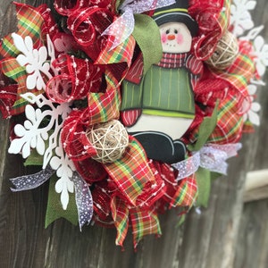 May include: A Christmas wreath with red and green plaid ribbon, red mesh, white snowflakes, and a snowman decoration.