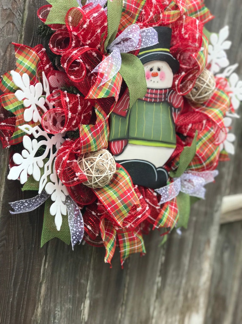 May include: A Christmas wreath with red and green plaid ribbon, red mesh, white snowflakes, and a snowman decoration.