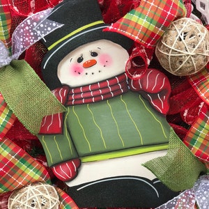 May include: A wooden snowman decoration with a black top hat, red and white striped scarf, and green sweater. The snowman is surrounded by red and green plaid ribbon, burlap, and white mesh.