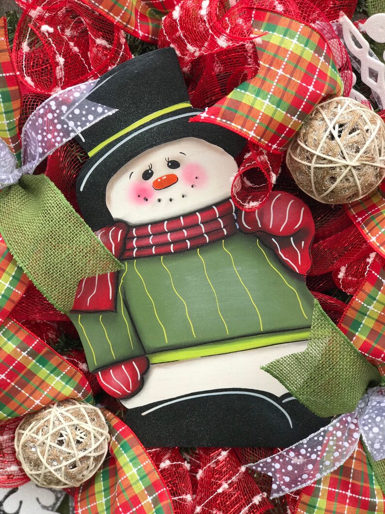 May include: A wooden snowman decoration with a black top hat, red and white striped scarf, and green sweater. The snowman is surrounded by red and green plaid ribbon, burlap, and white mesh.
