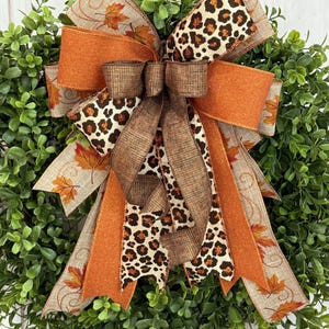 May include: A decorative bow featuring a mix of ribbons in fall colors. The bow includes ribbons with a leopard print, solid orange, and a beige ribbon with fall leaf patterns. The bow is set against a backdrop of green foliage.