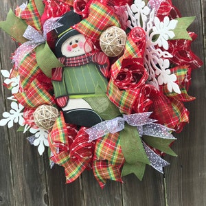 May include: A Christmas wreath with a snowman cutout, red and green plaid ribbon, white snowflake embellishments, and burlap accents.