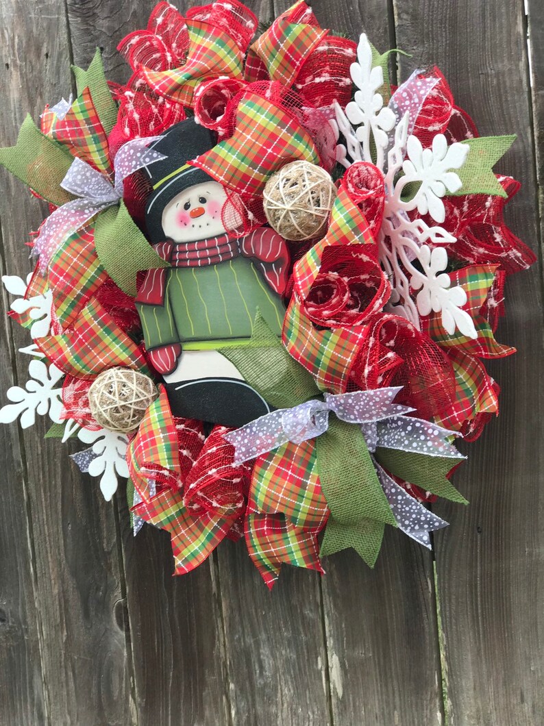 May include: A Christmas wreath with a snowman cutout, red and green plaid ribbon, white snowflake embellishments, and burlap accents.