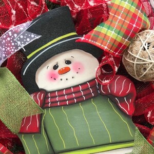 May include: A green and red snowman ornament with a black top hat and a white face. The snowman is surrounded by red and green plaid ribbon and burlap.