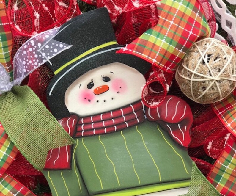 May include: A green and red snowman ornament with a black top hat and a white face. The snowman is surrounded by red and green plaid ribbon and burlap.