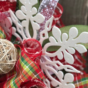 May include: Close-up of a white snowflake-shaped decoration on a Christmas wreath. The wreath is made of red and green plaid ribbon, red mesh, and a brown burlap ball.