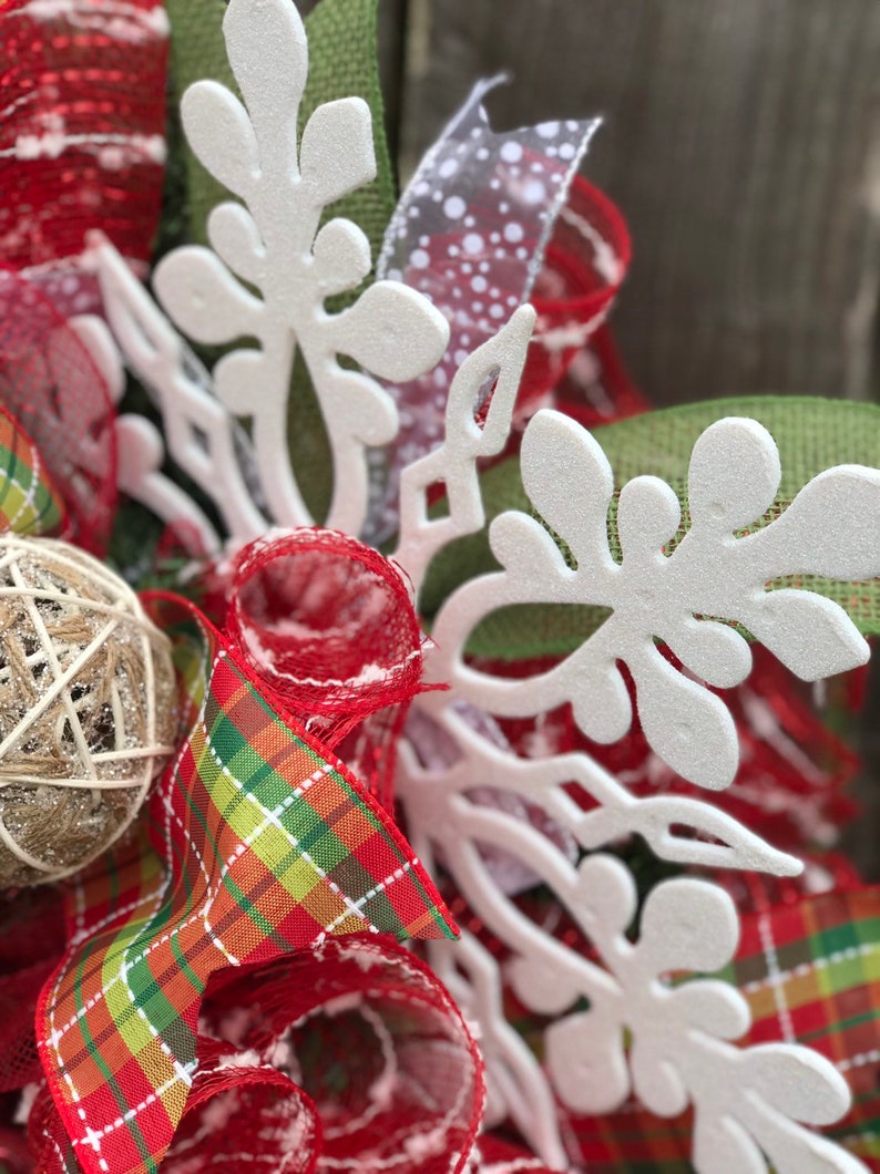May include: Close-up of a white snowflake-shaped decoration on a Christmas wreath. The wreath is made of red and green plaid ribbon, red mesh, and a brown burlap ball.
