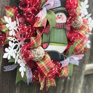 May include: A red, green, and white Christmas wreath with a snowman decoration. The wreath is made of mesh and ribbon and features white snowflakes.