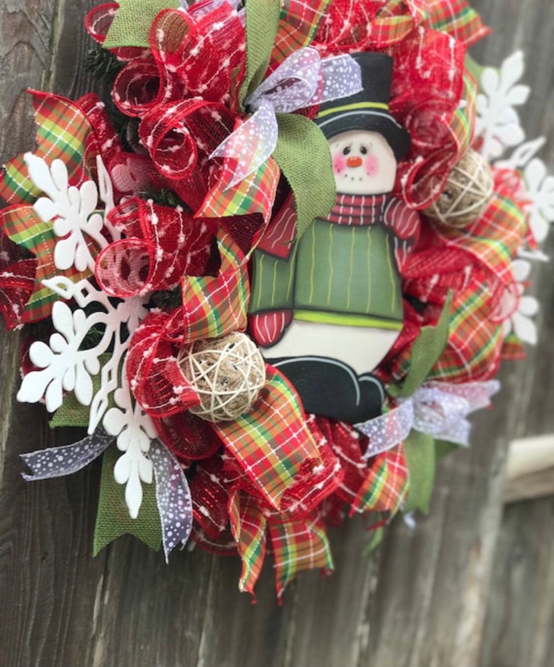 May include: A red, green, and white Christmas wreath with a snowman decoration. The wreath is made of mesh and ribbon and features white snowflakes.