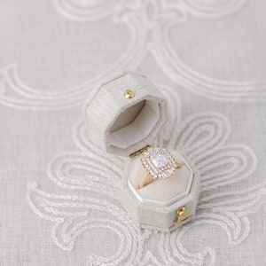 May include: A gold ring with a large, rectangular, clear diamond set in a halo of smaller diamonds. The ring is in a small, white, hexagonal box.