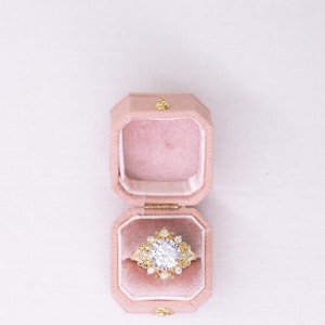 May include: A gold ring with a large, clear, round-cut centre stone and smaller surrounding stones, set in a pink velvet octagonal ring box. The ring box is open, revealing the ring's intricate design. The background is a soft white.