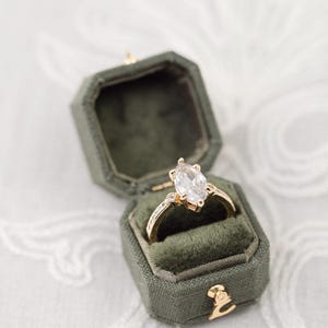 Könnte beinhalten: Ein goldener Verlobungsring mit einem großen, klaren, marquiseförmigen Edelstein wird in einer offenen, olivgrünen Samtringbox präsentiert. Die Ringbox hat einen goldenen Verschluss und befindet sich vor einem weißen, gemusterten Hintergrund.