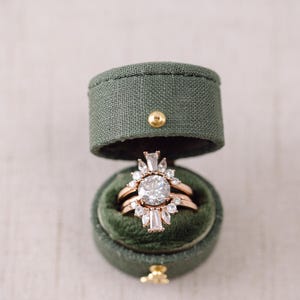 Puede incluir: Una caja de anillo con un interior de terciopelo verde y un anillo de oro con una piedra central grande y piedras más pequeñas alrededor.