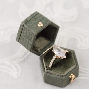May include: A gold engagement ring with a large, pear-shaped diamond is displayed in an open, olive-green velvet ring box. The ring box is hexagonal and has a gold clasp. The ring has smaller diamonds on the band.
