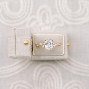 May include: A gold engagement ring with an oval-shaped diamond set in a delicate, floral-inspired band. The ring is displayed in a white velvet ring box.
