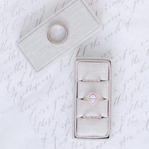 May include: An assortment of rose gold rings displayed in a light gray ring box and a separate ring box with a single ring. The rings include a band and two with small stones, and one with a large oval-shaped stone.