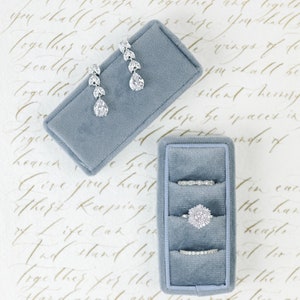 May include: A jewellery set comprising drop earrings and three rings, presented in a blue velvet box. The earrings and rings are embellished with clear, sparkling gemstones. The background features handwritten script.
