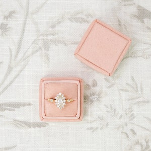 May include: A pear-shaped diamond engagement ring with a halo setting in a pink velvet ring box.