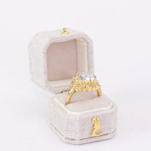 May include: A gold engagement ring with a large, clear center stone and smaller accent stones, set in a decorative setting. The ring is displayed in an open, light gray velvet ring box with gold accents.