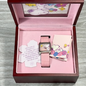 May include: A pink watch box featuring a white Snoopy character on the lid. Inside, a pink and silver watch with a Snoopy character on the face. The box also includes a certificate of authenticity confirming the watch is part of a limited edition of 2000.