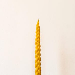 Puede incluir: Una vela alta de cera de abeja trenzada amarilla. La vela tiene un aspecto texturizado y tejido y se alza sobre una superficie de madera sobre un fondo blanco. La punta de la vela es de un tono amarillo más claro.