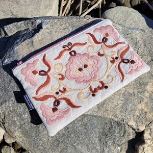 May include: A light pink linen pouch with a zipper closure and embroidered floral design in shades of pink, brown, and cream. The pouch is lying on a gray rock.